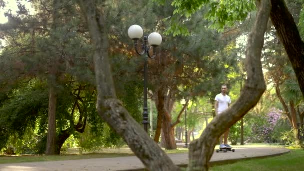 Jovem homem bonito em um capacete monta um skate elétrico em um belo parque de verão — Vídeo de Stock