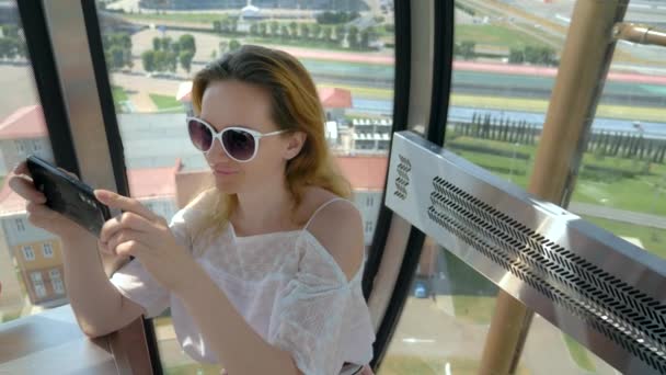 El concepto de ocio, viajes y entretenimiento. Una mujer hace una foto en su teléfono inteligente mientras disfruta de la vista desde la cabina de una rueda de hurón — Vídeos de Stock