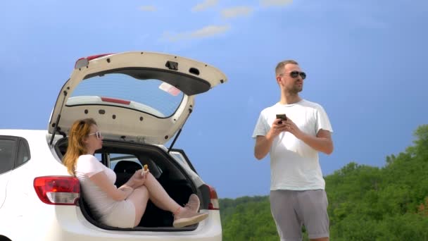 Viaje en coche y dos amantes hombre y mujer tomar un aperitivo mientras está sentado en el maletero de un coche contra el cielo azul y las montañas — Vídeo de stock