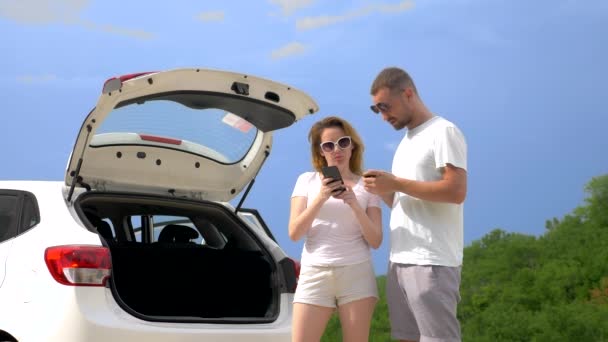 Konsept yol gezisi ve macera. bir çift mavi gökyüzü ve dağlara karşı arabanın yanında bir adam ve bir kadın gezinmek için akıllı telefonlarını kullanın — Stok video