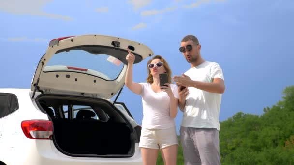 Concept road trip et aventure. un couple un homme et une femme près de la voiture contre le ciel bleu et les montagnes utilisent leurs smartphones pour naviguer — Video
