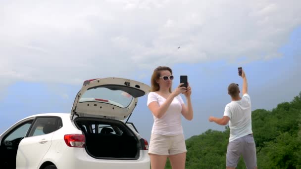 The concept of autotravel and adventure. couple man and woman near the car on a background of blue sky and mountains use their smartphones to find the signal of the cellular network — Stock Video