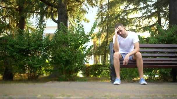 Депресивний сумний молодий чоловік сидить один на лавці в літньому парку. копіювання простору — стокове відео