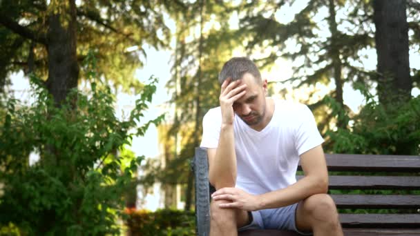 Depressieve trieste jonge man die alleen op een bankje in een zomerpark zit. ruimte kopiëren — Stockvideo