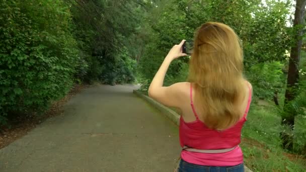 Фото или видео по телефону, женщина фотографирует на своем смартфоне. Летом красивый ландшафтный парк, парковая аллея — стоковое видео