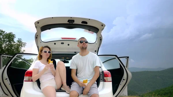 Viagem de carro e dois amantes homem e mulher ter um lanche enquanto sentado no porta-malas de um carro contra o céu azul e montanhas — Fotografia de Stock