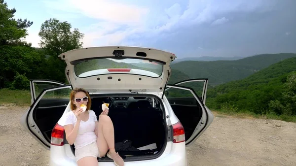 Uma viagem de carro e uma mulher para comer enquanto sentado no porta-malas do carro contra o céu azul e montanhas — Fotografia de Stock