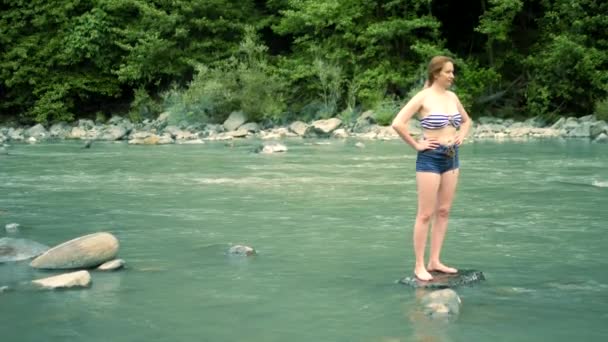Concetto di vacanza estiva. una donna in costume da bagno e pantaloncini va oltre un fiume di montagna a un guado — Video Stock