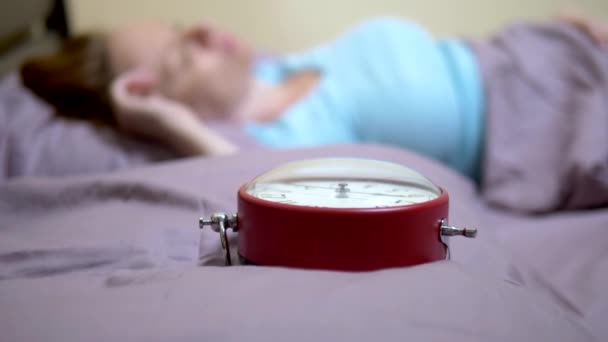 Крупним планом червоний будильник. На розмитому тлі дівчина в блакитній піжамі прокидається вранці — стокове відео