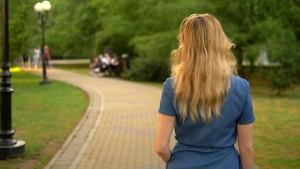 Вид сзади. средний план. красивая молодая блондинка в джинсовом комбинезоне прогуливается по летнему городскому парку — стоковое видео