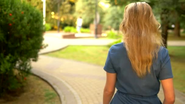Вид сзади. средний план. красивая молодая блондинка в джинсовом комбинезоне прогуливается по летнему городскому парку — стоковое видео