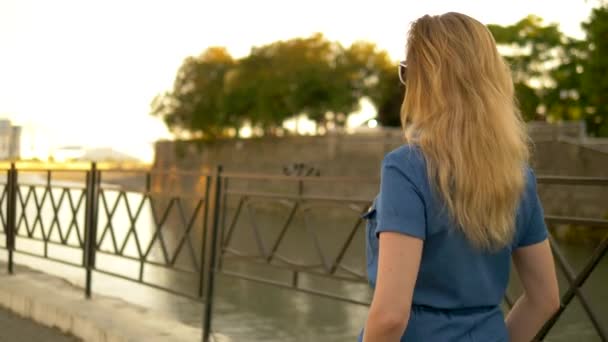 Vue arrière. plan moyen. belle jeune femme blonde en salopette denim se promène dans le parc de la ville d'été — Video