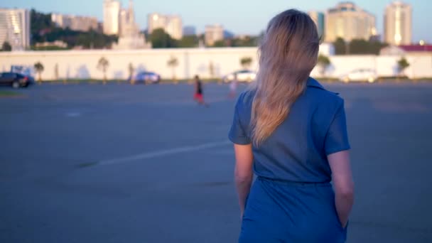 スタイリッシュなデニムのオーバーオールを着たスタイリッシュなブロンドの女性は、ヤシの木を背景に空の通りを歩いています。バックビュー — ストック動画