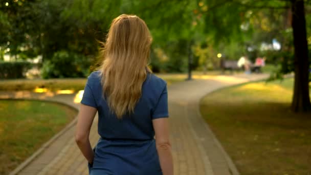 Vista posteriore. Piano medio. bella giovane donna bionda in tuta denim passeggiate attraverso il parco della città estiva — Video Stock