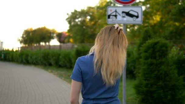 Vista posteriore. Piano medio. bella giovane donna bionda in tuta denim passeggiate attraverso il parco della città estiva — Video Stock