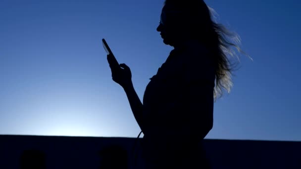 在蓝天日落的背景上，一个带着飞毛的智能手机的女人的剪影 — 图库视频影像