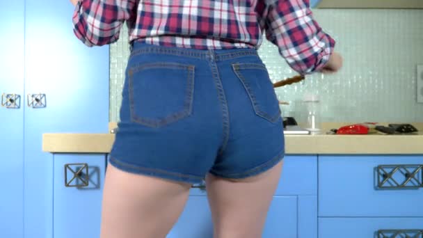 Sexig hemmafru förbereder mat på spisen hemma i korta jeansshorts och danser. lår närbild. — Stockvideo