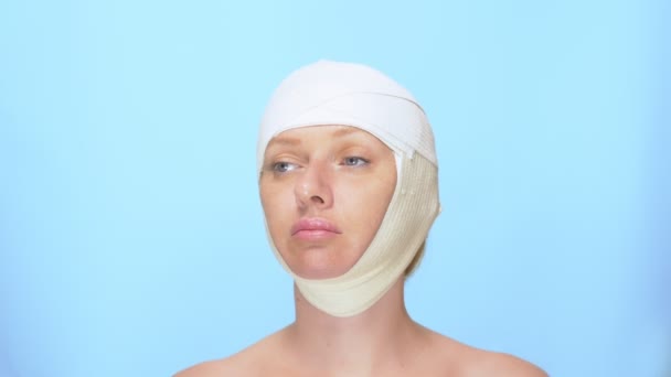 整形外科的概念。面部提升。一个头上有绷带的女人的肖像，在蓝色背景上. — 图库视频影像