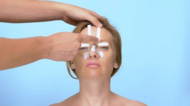 Plastik cerrahi kavramı. burun estetiği ve blefaroplasti. Doktorun elleri burnunda ve gözlerinde bandaj olan bir kadının yüzünü tutar. mavi arka planda