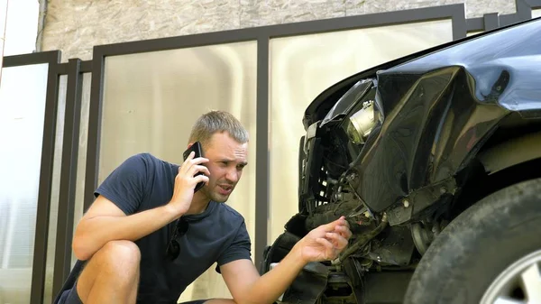 Car accident concept. man in a state of shock talking on the phone after a car accident, standing by a car with a broken bumper — Stock Photo, Image