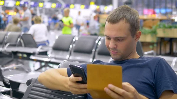 Bel homme à l'aéroport dans la salle d'attente. homme assis utilise un smartphone et détient un passeport avec des bagages . — Photo