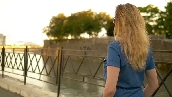 Back view. medium plan. beautiful young woman blonde in denim overalls walks through the summer city park — Stock Photo, Image