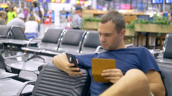 Красивый мужчина в аэропорту в приемной. Человек сидит использует смартфон и держит паспорт с багажом . — стоковое фото