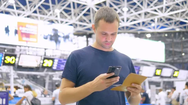 Un hombre guapo en el aeropuerto en la sala de espera. hombre se sienta utiliza un teléfono inteligente y tiene un pasaporte —  Fotos de Stock