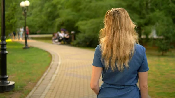 Vista posteriore. Piano medio. bella giovane donna bionda in tuta denim passeggiate attraverso il parco della città estiva — Foto Stock