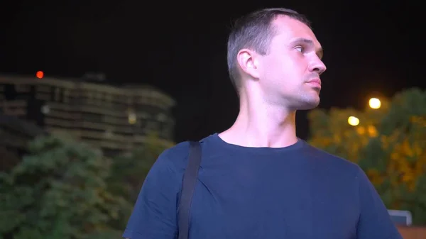Ein trauriger, gut aussehender Mann steht nachts auf der Straße. vor dem Hintergrund der nächtlichen Beleuchtung. schaut sich um und schaut auf dein Handy in Erwartung von etwas — Stockfoto
