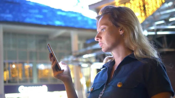 Eine traurige besorgte Frau steht nachts auf der Straße. vor dem Hintergrund der nächtlichen Beleuchtung. schaut sich um und schaut auf das Telefon in Erwartung von etwas — Stockfoto