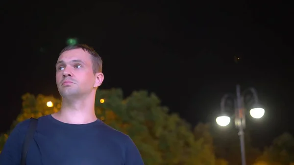 Ein trauriger, gut aussehender Mann steht nachts auf der Straße. vor dem Hintergrund der nächtlichen Beleuchtung. schaut sich um und schaut auf dein Handy in Erwartung von etwas — Stockfoto