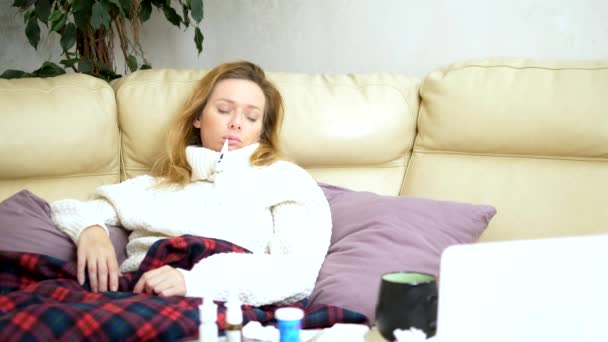 Ung sjuk kvinna i en tröja under en pläd med feber kontrollerar hennes temperatur med en termometer hemma — Stockvideo