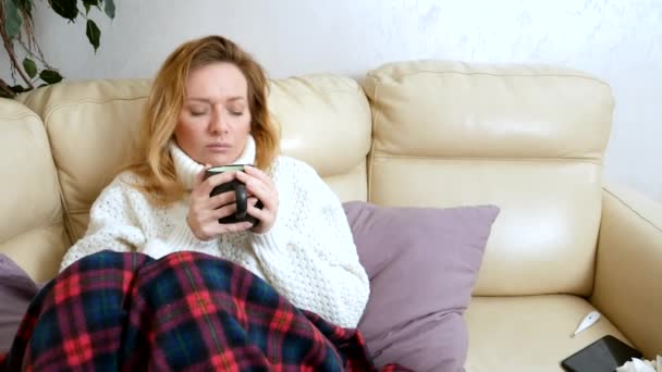 Zieke vrouw in een trui onder een deken het drinken van een warm drankje op de Bank in de woonkamer — Stockvideo