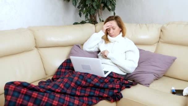 Blond woman with a cold sitting on the sofa in a sweater and a plaid calling her doctor on a laptop via video link. — Stock Video