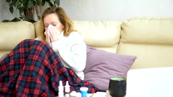 Λευκή γυναίκα, με χαρτί φτέρνισμα πετσέτα, σε ένα πουλόβερ κάτω από μια κουβέρτα στον καναπέ, βιώνει συμπτώματα αλλεργίας, πιάστηκε ένα κρύο. — Αρχείο Βίντεο