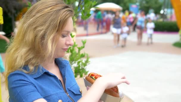 Giovane donna mangiare un hotdog appetitoso seduto nel parco sullo sfondo di persone che camminano, sfondo sfocato . — Video Stock