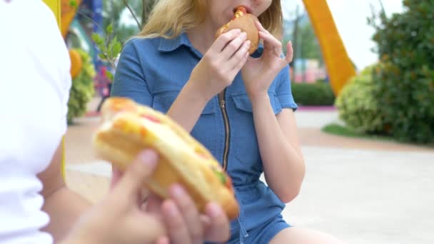 Pareja joven hombre y mujer comiendo un apetitoso perrito caliente, sentado en el parque contra el fondo de la gente que camina, fondo borroso . — Vídeo de stock