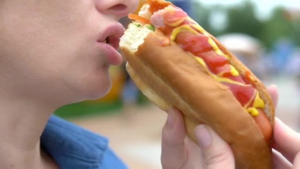 Zbliżenie. Młoda kobieta jedzenia apetyczny Hotdog siedzi w parku na tle chodzenia ludzi, niewyraźne tło. — Wideo stockowe