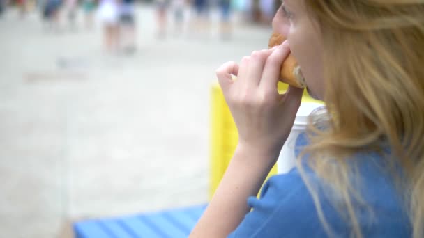 Primer plano. mujer joven comiendo un perrito caliente apetitoso sentado en el parque en el fondo de la gente que camina, fondo borroso . — Vídeos de Stock