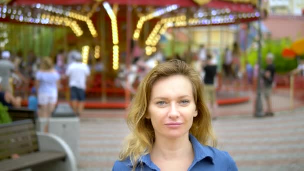 Концепция летнего времени. Женщина счастливо смотрит в камеру с доброй улыбкой на фоне карусели в парке развлечений — стоковое видео