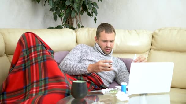 Homem com um frio sentado no sofá em uma camisola e uma xadrez chamando seu médico em um laptop via link de vídeo . — Vídeo de Stock