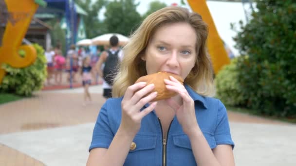 Mujer joven comiendo un perrito caliente apetitoso sentado en el parque en el fondo de la gente que camina, fondo borroso . — Vídeos de Stock