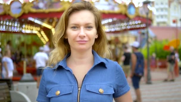 Concepto de hora de verano. Una mujer felizmente mira a la cámara con una amable sonrisa en el fondo del carrusel en un parque de diversiones — Vídeo de stock