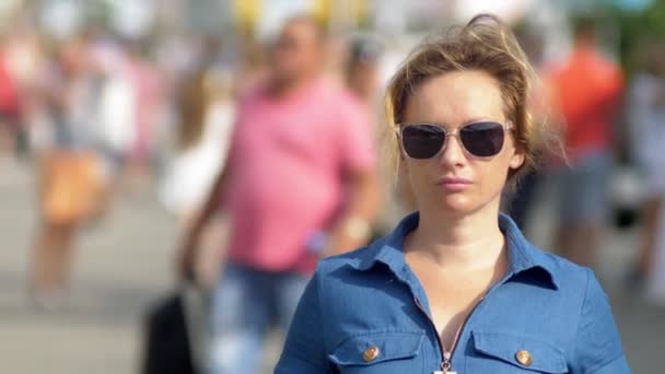 Hermosa mujer con gafas de sol, mirando a la cámara, de cerca. caluroso día de verano. Mucha gente en movimiento se desdibujó en el fondo . — Vídeo de stock