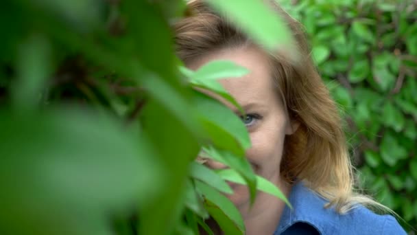 Een vrouw in een korte denim jumpsuit staande in het midden van een heggen doolhof op een zonnige zomerdag. Kijkt naar de camera. — Stockvideo