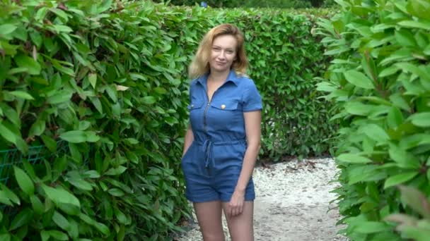 Een vrouw in een korte denim jumpsuit staande in het midden van een heggen doolhof op een zonnige zomerdag. Kijkt naar de camera. — Stockvideo