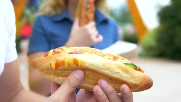 Koppel jonge man en vrouw eten een smakelijke hotdog, zittend in het Park tegen de achtergrond van het wandelen mensen, onscherpe achtergrond. — Stockvideo