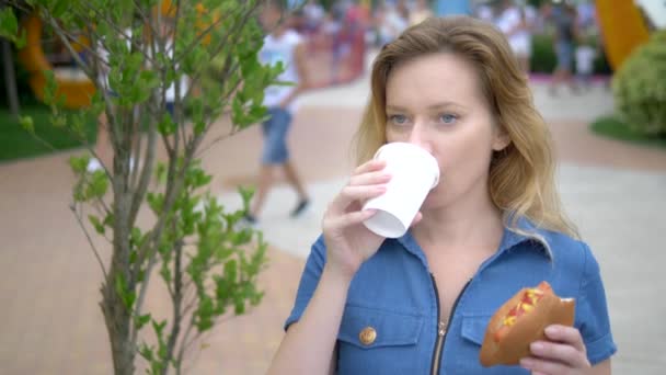 Fiatal nő eszik egy étvágygerjesztő hotdog ül a parkban a háttérben a gyaloglás emberek, homályos háttér. — Stock videók