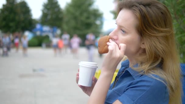 歩く人々の背景に公園に座って食欲をそそるホットドッグを食べている若い女性、ぼやけた背景. — ストック動画